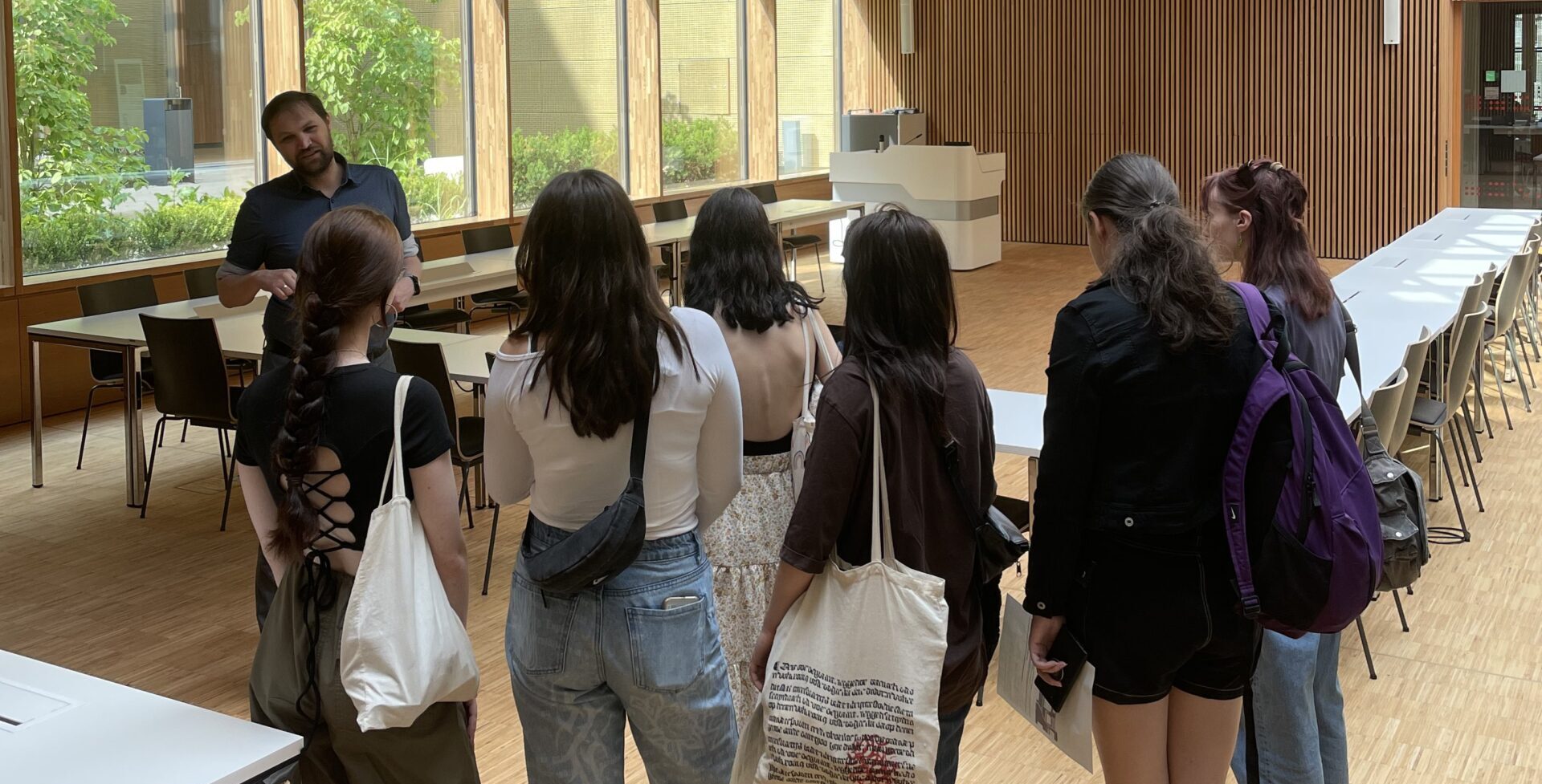 Eine Gruppe von Menschen steht in einem geräumigen, holzgetäfelten Raum mit großen Fenstern und Oberlicht in der Hochschule Coburg. Sie hören gespannt einem Redner am Tisch zu, während im Hintergrund ordentlich aufgestellte Tische und Stühle stehen.