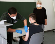 Eine Person mit Maske und Handschuhen verabreicht einer sitzenden Person an der Hochschule Coburg eine Impfstoffinjektion. Eine weitere Person steht in der Nähe und beobachtet den Vorgang. Alle sind maskiert und im Hintergrund ist eine grüne Tafel zu sehen.
