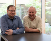 Zwei Männer, einer mit Brille und blauem Hemd, der andere mit hellem Hemd, lehnen lächelnd an einem dunklen Tisch im Innenbereich. Große Fenster geben den Blick auf üppiges Grün draußen frei. Die warme Atmosphäre lässt auf eine inspirierende Zusammenarbeit schließen, die möglicherweise mit der Hochschule Coburg in Verbindung steht.