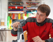 Ein junger Mann in rot-grauer Jacke sitzt lächelnd an einem Tisch und hält ein Werkzeug in der Hand. Hinter ihm sind bunte Stoffrollen und verschiedene andere Werkzeuge, die zweifellos Teil seiner kreativen Studienarbeit an der Hochschule Coburg sind. Der Raum gleicht einer lebendigen Werkstatt oder einem Atelier.