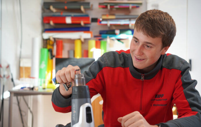 Ein junger Mann in rot-grauer Jacke sitzt lächelnd an einem Tisch und hält ein Werkzeug in der Hand. Hinter ihm sind bunte Stoffrollen und verschiedene andere Werkzeuge, die zweifellos Teil seiner kreativen Studienarbeit an der Hochschule Coburg sind. Der Raum gleicht einer lebendigen Werkstatt oder einem Atelier.