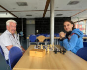 In einem modernen, hellen Raum der Hochschule Coburg sitzen zwei Personen an einem Tisch und spielen Schach auf einem Brett. Flankiert wird das Ganze von blauen Stühlen und hellen Holzmöbeln. Daneben steht eine Holzkiste. Einer von ihnen trägt eine blaue Jacke, der andere lächelt in einem hellen Hemd, während sie das Spiel spielen.