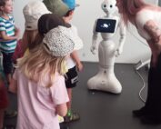 Eine Gruppe von Kindern und ein Erwachsener an der Hochschule Coburg beobachten einen humanoiden Roboter mit einem Bildschirm auf der Brust. Der Roboter steht in einem Raum mit einer schlichten Wand und die leger gekleideten Kinder wirken engagiert und neugierig.