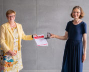 Zwei Frauen stehen vor einer grauen Wand an der Hochschule Coburg. Eine trägt eine gelbe Jacke und ein geblümtes Kleid und hält stolz eine rot-weiße Urkunde. Die andere Frau trägt ein marineblaues Kleid und zeigt eine glänzende Medaille. Beide lächeln strahlend.