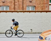 Eine Person mit Helm und Rucksack fährt mit dem Fahrrad auf einer Stadtstraße in der Nähe der Hochschule Coburg. Sie fahren an einer strukturierten weißen Wand vorbei, über der sich rote Backsteingebäude befinden, während im Vordergrund ein verschwommenes orangefarbenes Auto vorbeirast.