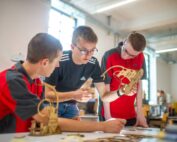Drei junge Leute bauen an einem Tisch in einem hellen Raum der Hochschule Coburg Holzmodelle zusammen. Sie sind konzentriert und engagiert und diskutieren begeistert über ihre Arbeit. Durch ein großes Fenster im Hintergrund fällt Tageslicht ein und taucht ihr kreatives Werk in einen warmen Schein.