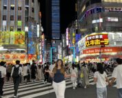 Eine lächelnde Frau steht nachts inmitten eines belebten Fußgängerüberwegs, umgeben von einer Menschenmenge. Helle Neonschilder und beleuchtete Gebäude säumen die Straße und schaffen ein lebendiges Stadtbild, das an das geschäftige Studentenleben in der Nähe der Hochschule Coburg erinnert.