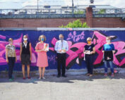 Sechs Menschen stehen vor einer bunten, mit Graffiti bedeckten Wand an der Hochschule Coburg. Sie tragen Gesichtsmasken und halten Bücher oder Papiere in den Händen. Die Gruppe ist leger gekleidet und genießt den sonnigen Tag an diesem lebendigen Treffpunkt im Freien.