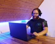 Eine Person mit Kopfhörern und Brille sitzt an einem Holztisch und beschäftigt sich mit einem Laptop. Vielleicht studiert sie Kurse der Hochschule Coburg. Der Raum mit seiner schrägen Holzdecke und dem blauen Umgebungslicht schafft eine ruhige Atmosphäre für konzentriertes Arbeiten.