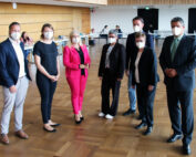 Sieben Personen stehen im Innenbereich der Hochschule Coburg, alle tragen Gesichtsmasken. Sie tragen eine Mischung aus formeller und halbformeller Kleidung. Einige andere sitzen an Tischen im Hintergrund, umgeben von dem Holzboden und den Holzwänden des Raums.