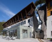 Modernes Gebäude mit Holz- und Glaselementen, an dessen Fassade sich eine abstrakte Glas- und Metallskulptur befindet. Vor einem klaren blauen Himmel und umgeben von Bäumen befindet sich dieses architektonische Juwel in der Nähe der Hochschule Coburg. Im Vordergrund befinden sich einladende Sitzbereiche im Freien.