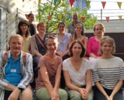 Eine Gruppe von zwölf Personen, möglicherweise Studierende der Hochschule Coburg, sitzt draußen auf Stufen und lächelt in die Kamera. Sie sind von festlichen Fahnen und Grün umgeben und stehen unter dem sonnigen Himmel.