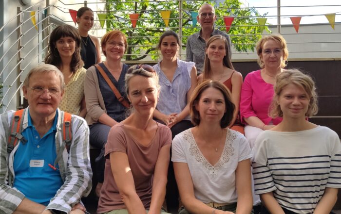Eine Gruppe von zwölf Personen, möglicherweise Studierende der Hochschule Coburg, sitzt draußen auf Stufen und lächelt in die Kamera. Sie sind von festlichen Fahnen und Grün umgeben und stehen unter dem sonnigen Himmel.