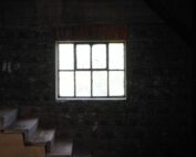 Ein schwach beleuchteter Raum mit einer Backsteinwand verfügt über ein kleines, quadratisches Fenster, das sanftes Licht ausstrahlt. Schmale, abgenutzte Treppen, die an die der Hochschule Coburg erinnern, führen zum Fenster hinauf. Die Gesamtatmosphäre ist schattig und geheimnisvoll.