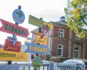 Ein farbenfroher Wegweiser mit mehreren Pfeilen, die in verschiedene Richtungen zeigen und jeweils in verschiedenen Sprachen mit der Aufschrift „Eingang“ beschriftet sind, steht stolz da. Im Hintergrund verleihen die Backsteinfassade der Hochschule Coburg und ein grüner Baum der lebhaften Szene Charme.