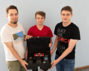 Drei junge Männer stehen zusammen und halten ein elektronisches Gerät mit einer digitalen Anzeige an der Hochschule Coburg. Das Gerät ähnelt einem Bedienfeld oder einem Puzzlespiel. Einer trägt ein weißes Hemd, ein anderer ein rotes und der dritte ein schwarzes Hemd mit Text. Sie lächeln in die Kamera und strahlen jugendliche Energie aus.