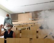 In einem Hörsaal der Hochschule Coburg führen drei Personen ein Experiment durch. Einer misst die Entfernung mit einem Maßband, ein anderer bedient die Ausrüstung und ein dritter sitzt inmitten von weißem Rauch oder Nebel. Alle tragen Masken, was darauf hindeutet, dass es sich bei dem Experiment um Luftströmungen oder Diffusion handelt.