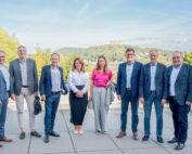 Eine Gruppe von acht Personen, fünf Männer und drei Frauen, posiert an einem sonnigen Tag im Freien an der Hochschule Coburg. Sie tragen Business-Kleidung. Hinter ihnen ist eine malerische Landschaft mit Hügeln und einem Schloss in der Ferne unter dem blauen Himmel zu sehen.