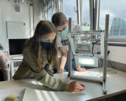 Zwei maskierte Studierende der Hochschule Coburg arbeiten fleißig in einem sonnendurchfluteten Klassenzimmer an einem Projekt mit einem Leuchtkasten und einem Metallrahmen; das Sonnenlicht strömt durch große Fenster.