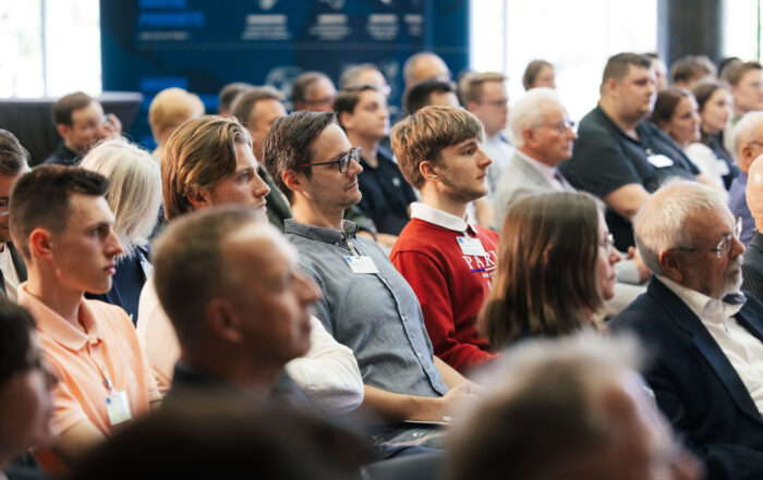 Eine bunt gemischte Gruppe von Menschen sitzt im Publikum und hört aufmerksam einer Präsentation an der Hochschule Coburg zu. Der Raum strahlt eine professionelle Konferenzatmosphäre aus, und ein blaues Banner im Hintergrund unterstreicht die akademische Atmosphäre.