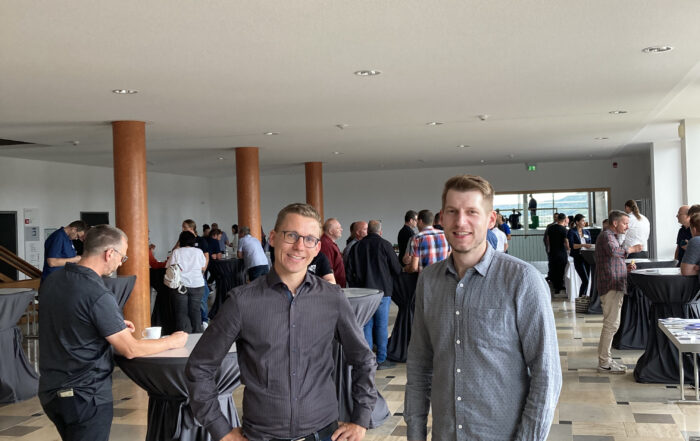 Zwei Männer stehen und lächeln in einem geräumigen Innenraum, vermutlich bei einer Networking-Veranstaltung der Hochschule Coburg. Im Hintergrund mischen sich mehrere Gruppen von Menschen und genießen die Atmosphäre, die durch große Fenster und hohe Holzsäulen entsteht.