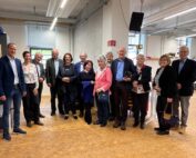 Eine Gruppe von zwölf Erwachsenen steht in einem Innenbereich der Hochschule Coburg auf einem Holzboden zusammen. Einige lächeln, sind leger gekleidet, im Hintergrund sorgen Regale und Decken im Industriestil für eine lockere und freundliche Atmosphäre.