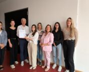 Eine Gruppe von acht Personen steht drinnen vor einer weißen Wand. Eine Person hält stolz ein Zertifikat der Hochschule Coburg in den Händen. Die bunt gemischte Gruppe, sowohl in Freizeit- als auch in Abendgarderobe gekleidet, strahlt lächelnd in die Kamera.