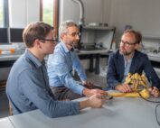 Drei Männer diskutieren und untersuchen in einem Labor an der Hochschule Coburg ein Skelettmodell auf einem Tisch. Sie sind konzentriert und in ein Gespräch vertieft. Im Hintergrund sind Computer und technische Geräte zu sehen, was ihr Engagement für die Weiterentwicklung des Wissens unterstreicht.