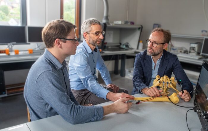 Drei Männer diskutieren und untersuchen in einem Labor an der Hochschule Coburg ein Skelettmodell auf einem Tisch. Sie sind konzentriert und in ein Gespräch vertieft. Im Hintergrund sind Computer und technische Geräte zu sehen, was ihr Engagement für die Weiterentwicklung des Wissens unterstreicht.