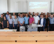 Etwa 35 Menschen, überwiegend Männer, haben sich in einem Hörsaal der Hochschule Coburg versammelt. Lässig gekleidet und lächelnd stehen sie vor einer Präsentationsleinwand. Im Raum stehen leere Holzbänke, vorne ist ein Projektor aufgebaut.