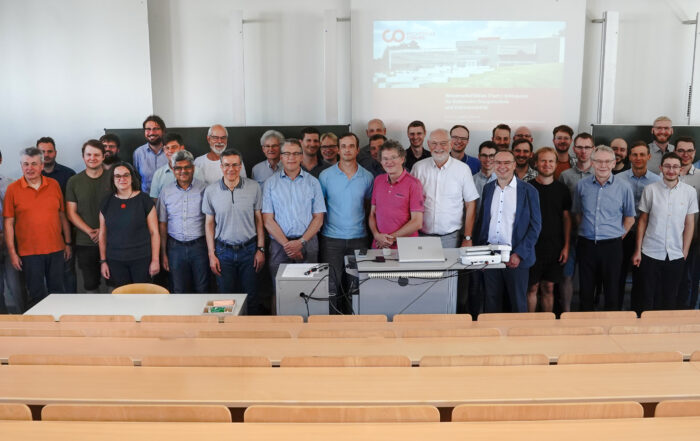 Etwa 35 Menschen, überwiegend Männer, haben sich in einem Hörsaal der Hochschule Coburg versammelt. Lässig gekleidet und lächelnd stehen sie vor einer Präsentationsleinwand. Im Raum stehen leere Holzbänke, vorne ist ein Projektor aufgebaut.