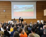 Eine große Gruppe von Menschen sitzt drinnen in der Hochschule Coburg vor einer Projektionsleinwand mit dem deutschen Text „HERZLICH WILLKOMMEN!“ Viele in der Menge haben ihre Hände gehoben. Der Raum hat holzgetäfelte Wände und eine sichtbare Uhr.