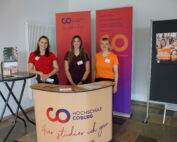 Drei Personen stehen bei einer geschäftigen Veranstaltung der Hochschule Coburg hinter einem Tresen. Der Tresen, geschmückt mit dem Logo und Branding der Hochschule, bietet informative Broschüren an. Im Hintergrund rücken ein Roll-Up-Banner und ein Informationstisch die Hochschule Coburg ins Rampenlicht.