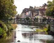 Eine schmale Brücke mit Blumenkästen überquert einen ruhigen Bach in einer malerischen Stadt in der Nähe der Hochschule Coburg. Zwei Menschen und ein Hund gehen über die Brücke. Unter dem hellen Himmel sind von Bäumen gesäumte Straßen und charmante Häuser mit Ziegeldächern zu sehen.