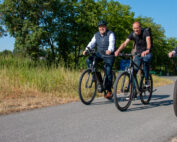 Drei Männer der Hochschule Coburg fahren mit dem Fahrrad auf einem gepflasterten Weg inmitten von Grün. Sie lächeln, tragen legere Kleidung und Helme und genießen den sonnigen Tag unter dem klaren blauen Himmel.