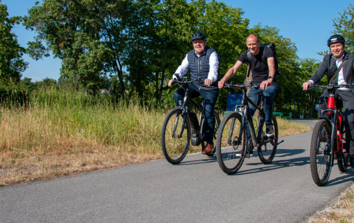 Drei Männer der Hochschule Coburg fahren mit dem Fahrrad auf einem gepflasterten Weg inmitten von Grün. Sie lächeln, tragen legere Kleidung und Helme und genießen den sonnigen Tag unter dem klaren blauen Himmel.