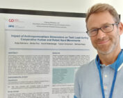 Eine Person mit Brille steht neben einem wissenschaftlichen Poster mit dem Titel „Einfluss anthropomorphistischer Dimensionen auf die Aufgabenbelastung bei kooperativen menschlichen und Roboterhandbewegungen“ in einem Robotiklabor der Hochschule Coburg. Das Poster enthält Abschnitte zu Zusammenfassung, Studie und Ergebnissen.