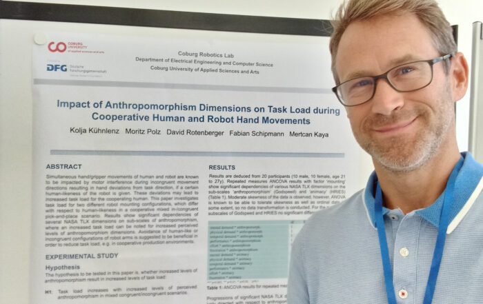 Eine Person mit Brille steht neben einem wissenschaftlichen Poster mit dem Titel „Einfluss anthropomorphistischer Dimensionen auf die Aufgabenbelastung bei kooperativen menschlichen und Roboterhandbewegungen“ in einem Robotiklabor der Hochschule Coburg. Das Poster enthält Abschnitte zu Zusammenfassung, Studie und Ergebnissen.