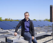 Eine Person sitzt auf einer Metallkonstruktion neben Solarmodulen unter einem klaren blauen Himmel, vermutlich an der Hochschule Coburg. Sie trägt eine schwarze Jacke und ein blaues Hemd. Im Hintergrund sind Bäume und ein Schornstein zu sehen.