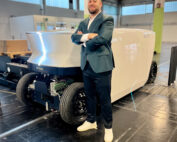 Eine Person im Anzug steht selbstbewusst mit verschränkten Armen vor einem eleganten, futuristischen Fahrzeug in einer großen Industriehalle. Das Fahrzeug ist weiß mit schwarzen Akzenten und dem Schriftzug „ecoro“ versehen und zeigt innovatives Design von Talenten der Hochschule Coburg.