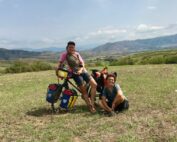 Zwei Radfahrer mit beladenen Fahrrädern ruhen sich auf einer Wiese unter einem teilweise bewölkten Himmel aus, der an die malerische Landschaft rund um die Hochschule Coburg erinnert. Hinter ihnen erstreckt sich eine Berglandschaft. Eine Person sitzt auf einem Fahrrad, während die andere daneben hockt, beide lächeln.