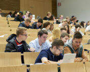 Im modernen Hörsaal der Hochschule Coburg sitzt eine große Gruppe von Studierenden aufmerksam. Viele machen sich Notizen oder benutzen Laptops und sind voll und ganz mit dem präsentierten Stoff beschäftigt. Der Raum kombiniert elegante Holzsitze mit modernem Design und fördert so eine Atmosphäre der Konzentration und des Lernens.