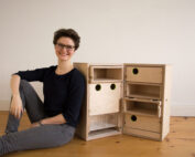 Eine Person mit kurzen Haaren und Brille sitzt auf einem Holzboden neben einem einzigartigen Schrank, der mit kreisrunden grünen Löchern verziert ist. Die offenen Fächer des Schranks offenbaren sein faszinierendes Design, das an den kreativen Geist der Hochschule Coburg erinnert. Eine schlichte, helle Wand vervollständigt die ruhige Szene.