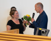 Ein Mann und eine Frau unterhalten sich mit Mikrofonen an der Hochschule Coburg. Die Frau mit einem Blumenstrauß in der Hand steht neben ihm in einem Raum, im Vordergrund sind ein Rednerpult und ein Projektor zu sehen. Im Hintergrund ist eine weitere Person unscharf zu sehen.