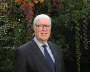 Ein Mann mit weißem Haar und Brille, einem dunklen Anzug und einer blauen Krawatte, lächelt herzlich vor einer Kulisse aus üppigem Grün mit roten und grünen Blättern und verkörpert den vornehmen Charme der Hochschule Coburg.
