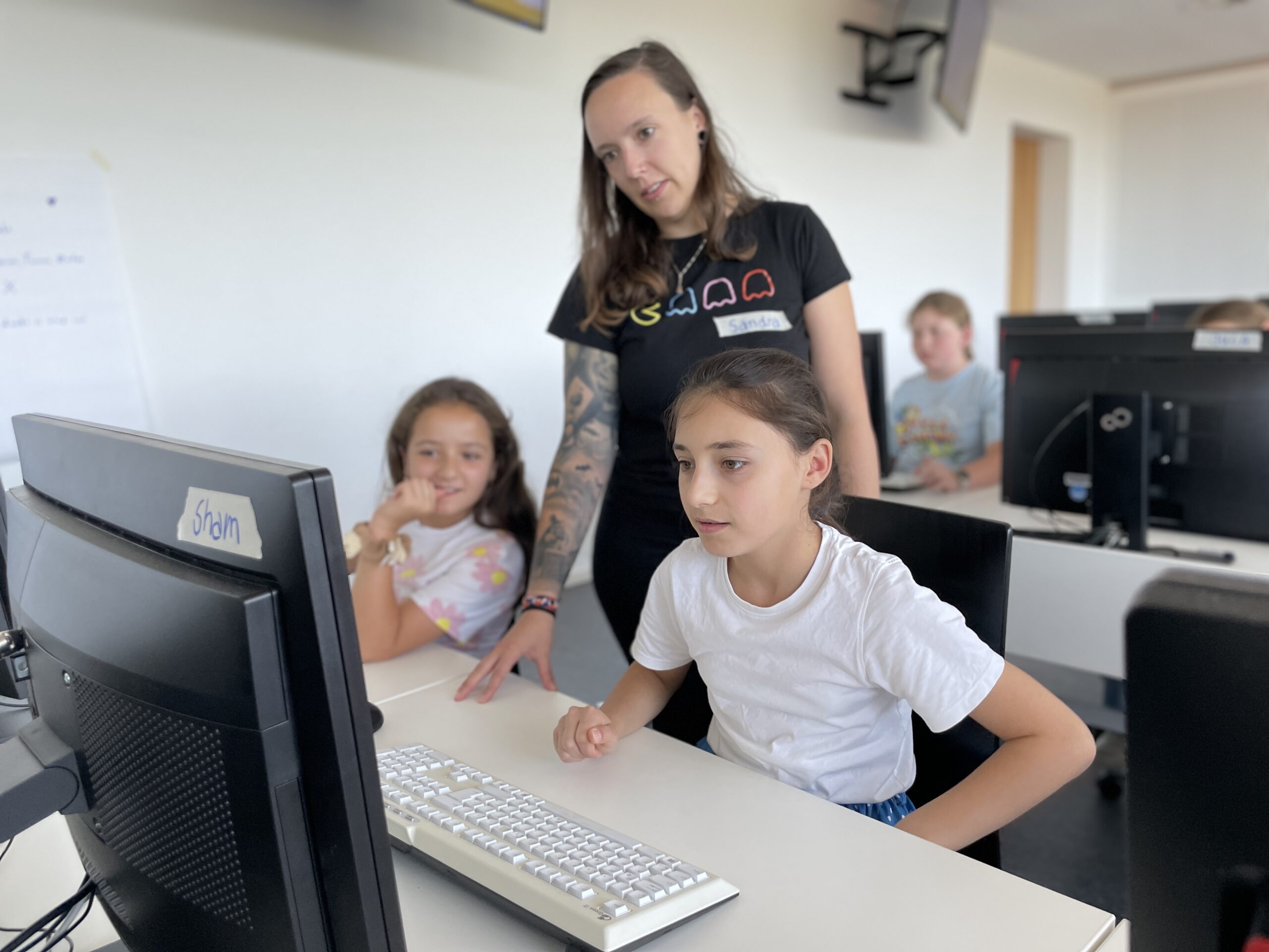 Ein Lehrer steht neben zwei jungen Mädchen, die an Computertischen sitzen und auf einen Bildschirm blicken. Die Schülerinnen beschäftigen sich mit dem Computer, während andere im Hintergrund stehen. Die Umgebung scheint ein Klassenzimmer oder eine Werkstatt der Hochschule Coburg zu sein, in der technologische Fähigkeiten vermittelt werden.