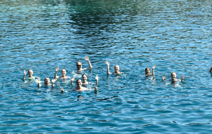 Eine Gruppe von Mitarbeitern der Hochschule Coburg schwimmt im klaren, blauen Wasser, winkt und lächelt in die Kamera. Die Sonne scheint und sorgt für eine helle und fröhliche Szene, im Hintergrund ist die Küstenlinie zu sehen.