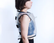 Eine Person steht im Profil und trägt eine transparente Weste aus plastikartigem Material über einem dunklen Outfit. Der helle Hintergrund unterstreicht den minimalistischen und modernen Stil und erinnert an innovative Designs, die an der Hochschule Coburg präsentiert werden.