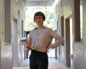 Eine Person mit kurzen Haaren und Brille steht in einem Flur der Hochschule Coburg und hält mehrere Bücher und Papiere in der Hand. Der Flur ist modern gestaltet und hat auf beiden Seiten symmetrische Öffnungen, während am Ende Sonnenlicht durch das Glas fällt.