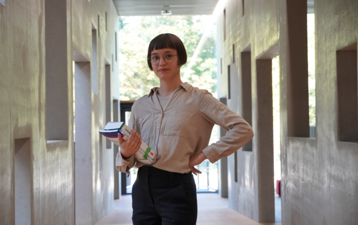 Eine Person mit kurzen Haaren und Brille steht in einem Flur der Hochschule Coburg und hält mehrere Bücher und Papiere in der Hand. Der Flur ist modern gestaltet und hat auf beiden Seiten symmetrische Öffnungen, während am Ende Sonnenlicht durch das Glas fällt.